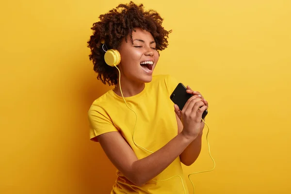 Joyful optimistic glad black woman glad to search favorite composition on web page, listens favorite cool song in headphones, sings from pleasure, has modern gadgets, wears yellow t shirt.
