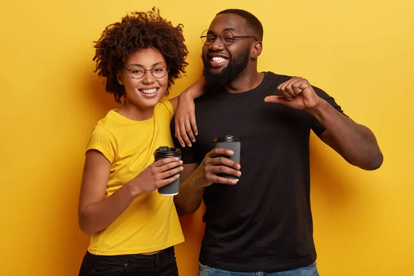 Happy black male student proud of himself as passed exam successfully, drinks takeaway coffee with female groupmate, enjoy spare time after classes, have hot drink, isolated over yellow wall