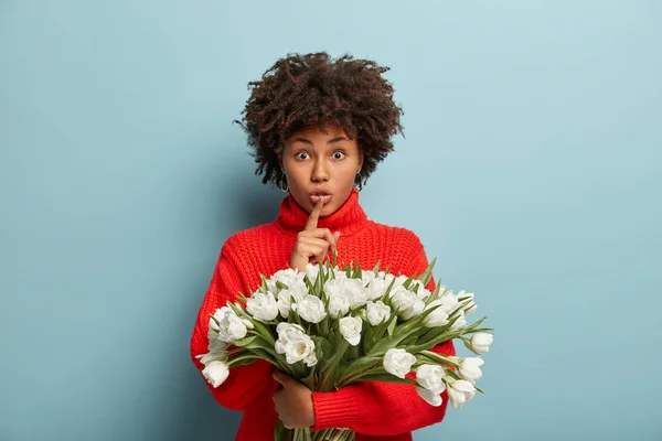 Photo of secret woman makes quiet gesture, keeps index finger over lips, looks surprisingly, carries huge bouquet of tulips, wants to make surprise for mother, isolated over blue wall.