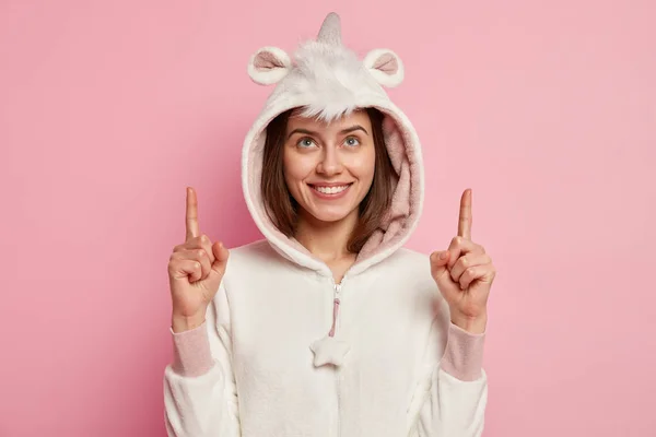 Optimistic lovely woman with satisfied expression, points above, wears domestic costume with hoody and animal ears, shows best copy space you ever seen, smiles friendly at camera, being at home