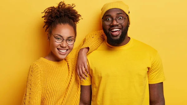 Photo of cheerful dark skinned young lady and man stand closely, enjoy spare time, listens funny news from interlocutor, wear spectacles and yellow clothes.