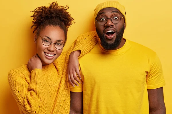 Emotional surprised black young man opens mouth from surprise, keeps hand on neck, has curly hairstyle, wears round spectacles, isolated over yellow studio. Monochrome.