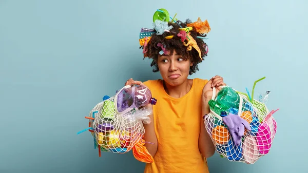 Confused hesitant dark skinned lady looks with questioned puzzled expression aside, doesn\'t know what to do with collected trash, busy cleaning during World Environment Day.