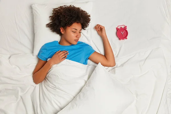 Cute restful lady with Afro haircut, lies under white blanket, has deep healthy sleep, has much time before awakening, poses in bed with alarm clock near, photographed from above with copy space