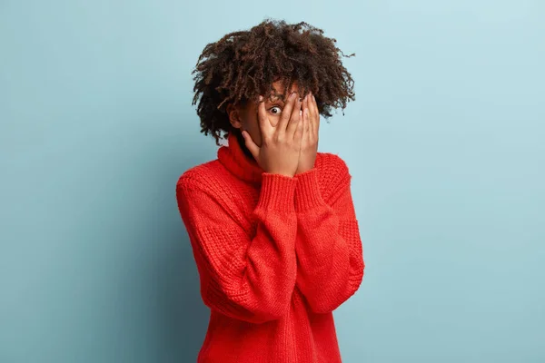 Indoor shot of young lady with Afro hair covers face with hands, peeks through fingers, hides from someone, has surprised expression wears red clothing afraid of something near. Oh no, take this away