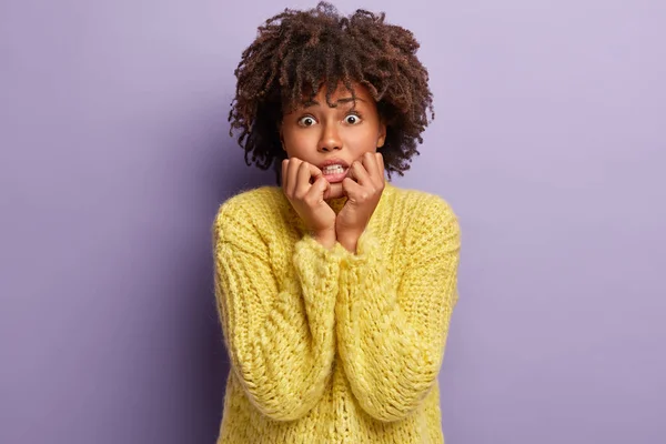 Image of nervous dark skinned female bites finger nails from depression, worries because of hurt feelings and separation with boyfriend, has Afro haircut, wears yellow jumper, poses indoor alone