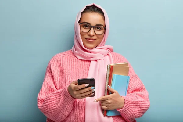 Horizontal shot of satisfied college student uses new cool app on cell phone, carries notepad for writing notes, wears spectacles, silk scarf and knitted sweater, isolated over blue background.