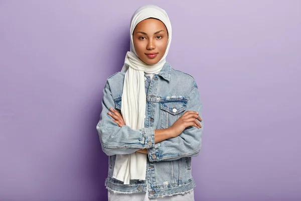 Attractive Eastern woman covers head with white headscarf to guard her dignity and power, has special dress code, keeps hands crossed, looks with modesty, poses over purple background.