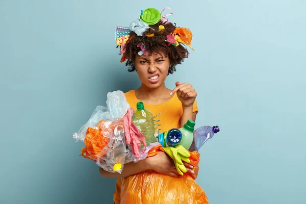 Voluntary free assistance. Irritated black woman worried with environmental pollution problem, carries trash bag with recycled plastics, clenches teeth from annoyance, isolated over blue studio wall