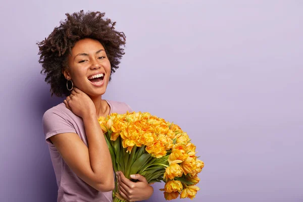 Sideways shot of glad dark skinned woman laughs with joy, touches neck, holds yellow tulips, wears violet t shirt, pleased by getting flowers and compliment, poses over purple studio wall, free space