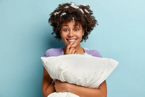 Indoor shot of glad dark skinned woman holds soft white pillow, sleeps well at night, enjoys rest, comfort and leisure, has cheerful expression, stands over blue background.