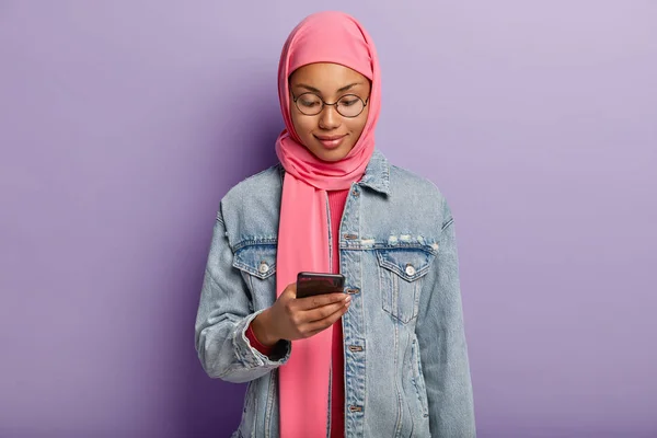 Horizontal shot of concentrated delighted dark skinned female of Muslim religion, types messages on modern cell phone, wears hijab and denim jacket, reads notification, surfs wireless internet