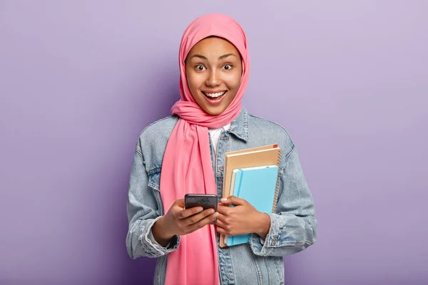 Happy Muslim woman advanced user of technology holds notepads and cellular, wears pink veil on head, fashionable denim jacket, reads new message in social networks, glad to have money on balance