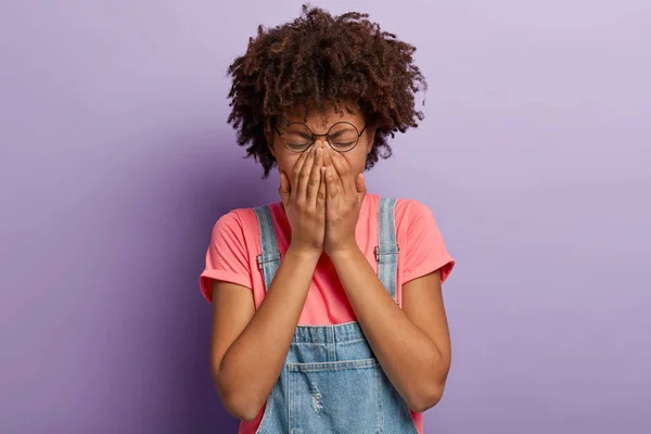 Discouraged desperate woman covers face, whines from negative news, has sad facial expression, wears round spectacles, t shirt and denim overalls, disappointed by something bad, poses in purple studio