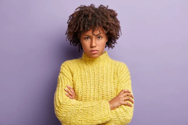 Photo of self confident serious woman with Afro hair, keeps arms folded, looks angrily at camera, wears winter yellow sweater, stands against purple background.