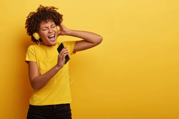 Pleased stylish dark skinned girl enjoys music from motivation playlist, enjoys free time for listening popular tracks, sings loudly words of song, closes eyes from satisfaction, wears yellow t shirt