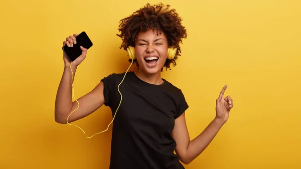 Funny dark skinned female feels great, dances to rhythm, shakes raised hands, sings along with music, wears headphones, holds modern cellular, dressed in t shirt, models over yellow background