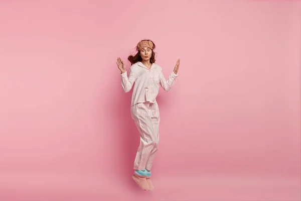 Horizontal shot of funny woman dressed in domestic clothing, has fun before sleep, jumps in air over pink background. Pretty lady wears pyjamas and sleepmask, photographed in motion, has lips folded