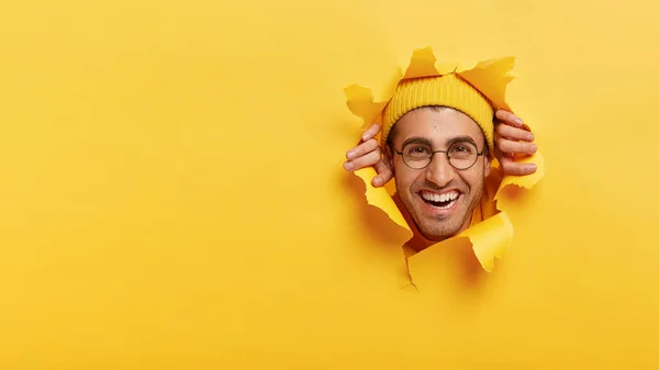 Joyful satisfied young Caucasian man wears optical glasses, yellow headgear, looks glad fully through hole in paper yellow wall, empty space for your advertising content, promotion or slogan.