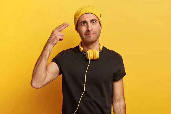 Horizontal shot of Caucasian man with dark hair, shoots in temple, demonstrates suicide gesture, tired from his failures, listens music from playlist in headphones, wears yellow hat and black t shirt