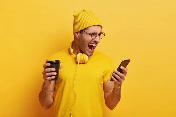 Irritated guy looks angrily at smart phone, receives unpleasant message, screams negatively, wears yellow stylish outfit, round spectacles drinks coffee to go, stands in studio.