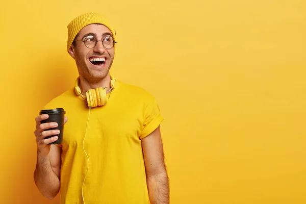 Optimistic man in glasses laughs as spends time with friends during coffee break, holds disposable cup, looks away, wears stylish yellow outfit, listens music in headphones, enjoys nice weekend