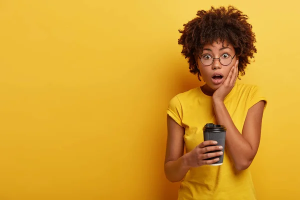 Photo of surprised dark skinned woman looks in stupor at camera, keeps hand on cheek, opens mouth widely, dressed in casual yellow t shirt, drinks coffee to go.