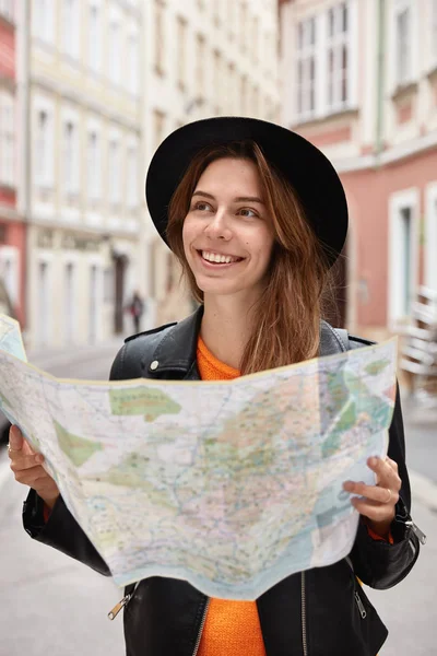 Happy holidaymaker searches right direction on map, explores new town with many sights, thinks about next tour, wears hat and black leather coat, smiles happily.