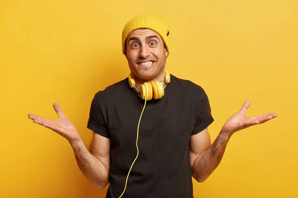 Horizontal shot of optimistic Caucasian man spreads hands in different sides, has surprised cheerful expression, wears yellow hat and casual t shirt, headphones on neck connected to some device