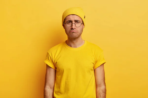 Unhappy unsatisfied guy smirks face, has gloomy expression, wears yellow hat and t shirt, puzzled by bad news, poses over studio wall, being offended by someone.