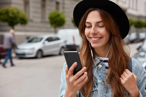 Pleased female in hat, holds cell phone in hand, listens music from playlist, strolls during free time over urban setting, has spring walk outdoor, copy space aside for your advertising content, chats