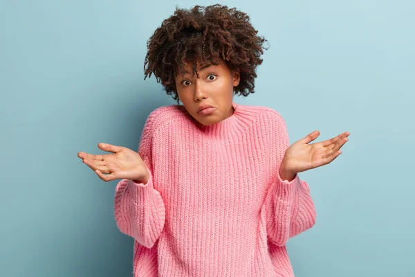 Doubtful African American woman spreads hands, feels uncertain, faces unfamiliar topic, feels perplexed while tries to find answer, wears pink oversized jumper, holds hands sidewards, has no idea