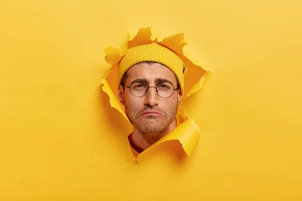 Gloomy dissatisfied Caucasian man smirks face from negative emotions, has sad look, wears yellow hat, round transparent glasses, looks through torn paper hole, isolated over yellow background