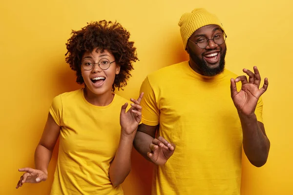 Photo of happy African couple dance together against yellow background, move body actively, show okay gesture, wear casual yellow t shirt, have fun during party. Monochrome. Feeling rhythm of music