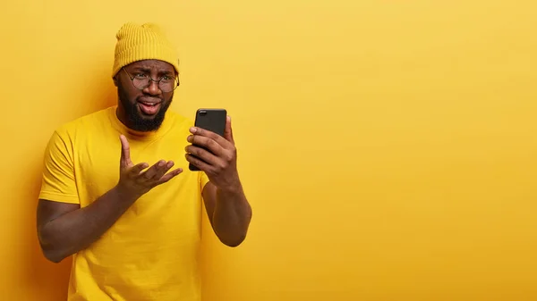 Devastated dissatisfied man with dark skin, gestures and looks at smart phone device, has problem with application, has thick bristle, wears round spectacles and casual t shirt, isolated on yellow