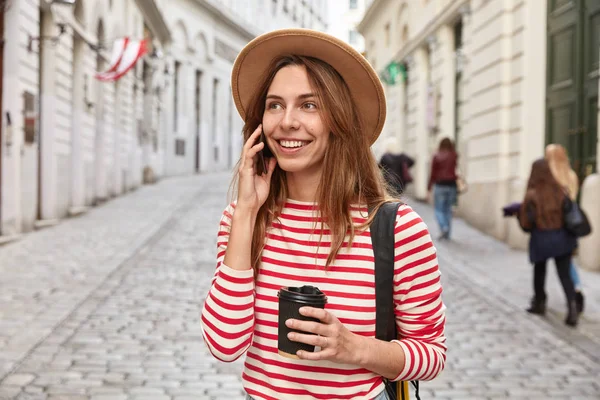 Gorgeous smiling tourist strolls around city, has phone conversation, holds takeaway coffee, focused somewhere into distance, wears stylish apparel, uses cheap tariffs, travels around world.