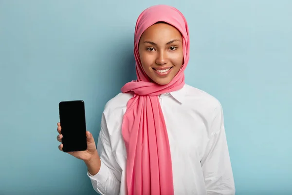 Beautiful Muslim woman advertises modern gadget, holds smart phone device with blank screen for your advertisement, wears traditional veil on head, white shirt, isolated on blue wall, smiles gently