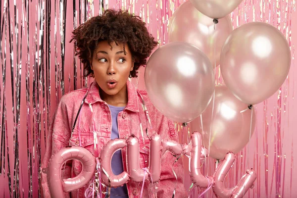 Photo of impressed dark skinned woman with make up looks surprisingly aside, being speechless, has bated breath, holds balloons, has tinsel on body, isolated over pink background.