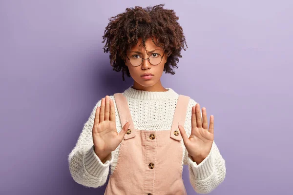 Dissatisfied young woman with dark skin, curly hair, makes stop gesture, has angry facial expression, shows palms at camera, denies participating in something, stands indoor. No sign and rejection
