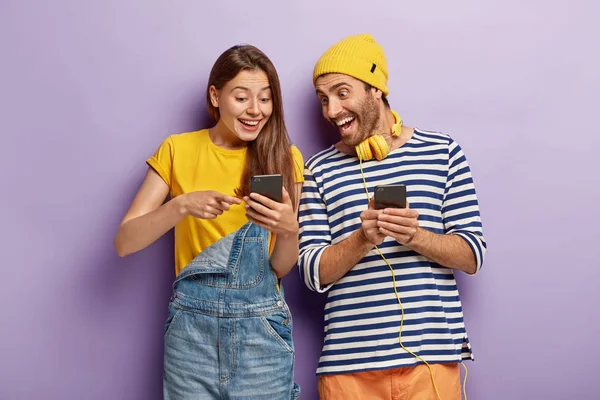 Horizontal shot of optimistic joyful couple chat with followers from web blog, gaze happily at smart phone, wear fashionable clothes, stand indoor.