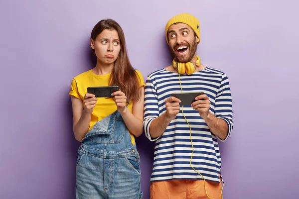 Happy young man and sad female bloggers use smartphone devices for online communication, play games, being addicted to modern technologies, stand shoulder to shoulder, express different emotions