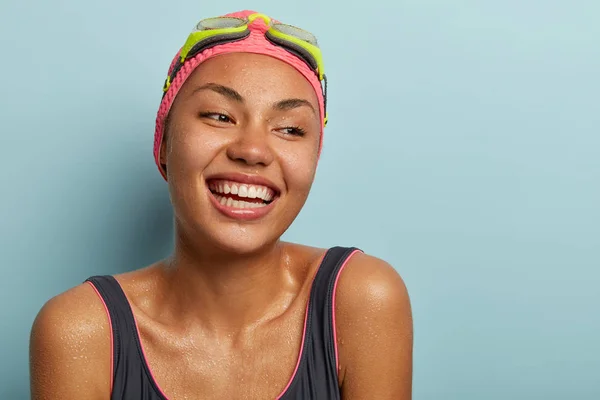 Close up portrait of relaxed happy dark skinned lady feels refreshed after active swim, has water dopes on smooth skin, satisfied to have summer rest at awesome place, poses in black swimsuit