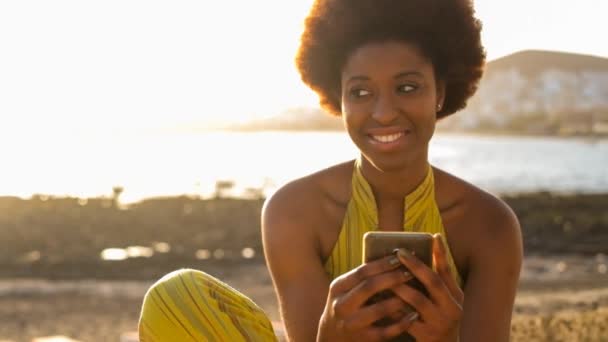 美丽的黑色种族非洲模式与漂亮的头发使用智能手机坐在附近的海洋和海滩自由休闲度假活动 Wifi 与朋友或父母联系和连接 — 图库视频影像