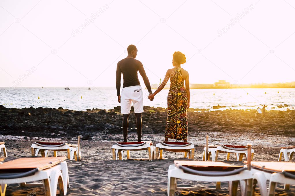 very romantic black young guys model couple african race hands by hands stand up looking themselves with a sunset in background. sand beach and seats closed for an infinity love concept