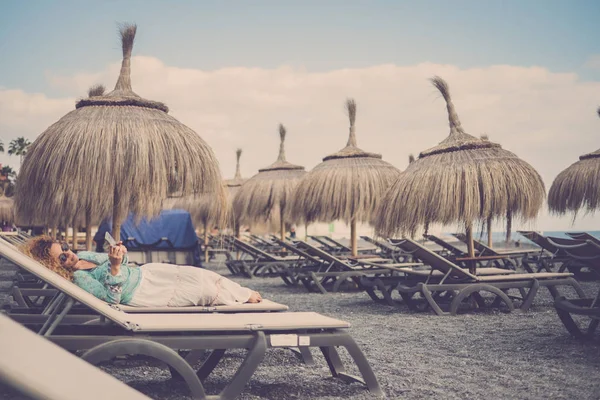 beautiful caucasian female middle age lay down on the seats at the beach with tropical umbrellas. travel and vacation rest concept. stay connected with mobile phone to chat with friends or parents using internet