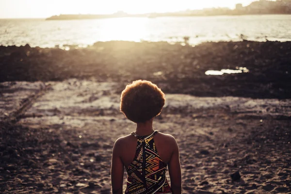 black race model with nice hair viewed from the back with rocky beach and ocean sunset light golde background. freedom and wanderlust concept for traveler and people who love the world