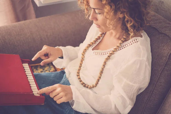 beautiful caucasian curly hair model middle age woman at home playing a little piano toy. artist at work sitting on the sofa. lonely female study music alone for hobby or work