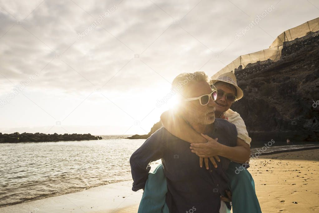 Happy senior adult caucasian couple in love. the man carry on his back the beautiful woman and both smiles. effective and together forever life concept in vacation time , sunset backlight and ocean waves
