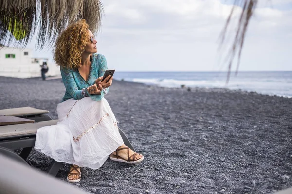 Sahilde okyanus ve dalgalar onay sırasında belgili tanımlık smartphone ile internet bakarak siyah volkanik bir koltukta oturan güzel beyaz bayan. yaşam tarzı alternatif iş ya da arkadaşlarınızla bağlamak