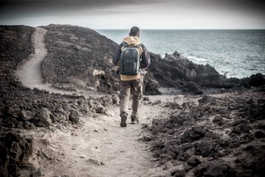 trekking giysi ve bir taş çölde Okyanusu kıyı şeridi üzerinde yürüyen sırt çantası ile yalnız seyahat adam genç orta yaş. deniz manzaralı bir yolunu izleyerek ve özgürlük ve alternatif yaşam tarzı zevk